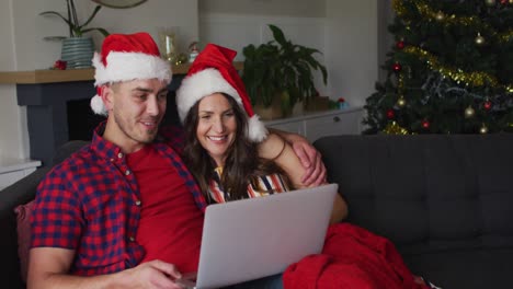 Glückliches-Kaukasisches-Paar-Mit-Weihnachtsmützen,-Das-Auf-Dem-Sofa-Sitzt-Und-Einen-Laptop-Benutzt