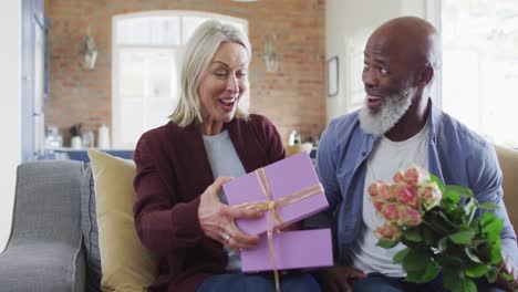 Happy-senior-diverse-couple-in-living-room-sitting-on-sofa,-giving-flowers-and-present