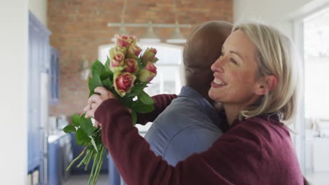 Happy-senior-diverse-couple-in-living-room-embracing,-holding-flowers