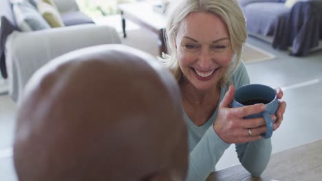 Caucasian-senior-woman-holding-a-coffee-cup-smiling-while-talking-to-her-husband-at-home