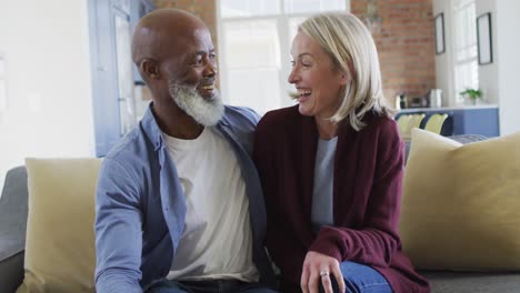 Portrait-of-happy-senior-diverse-couple-in-living-room-sitting-on-sofa,-embracing-and-smiling