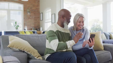 Happy-senior-diverse-couple-in-living-room-sitting-on-sofa,-using-smartphone,-making-video-call