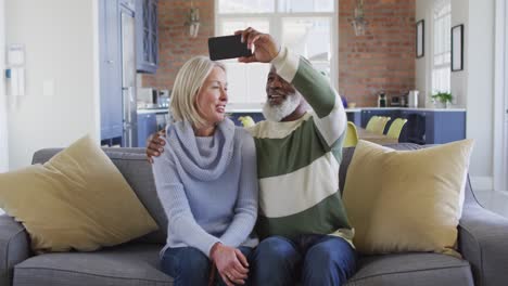 Happy-senior-diverse-couple-in-living-room-sitting-on-sofa,-using-smartphone,-making-video-call