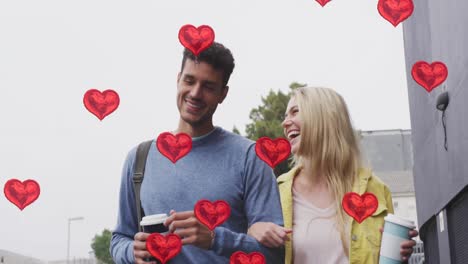 Animation-of-heart-icons-floating-over-happy-caucasian-couple-walking-and-drinking-takeaway-coffee