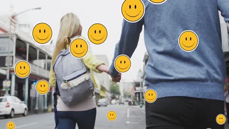 Animation-of-digital-emoji-icons-floating-over-happy-caucasian-couple-walking-and-holding-hands