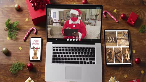 African-american-santa-in-face-mask-on-video-call-with-smartphone,-tablet-and-christmas-decorations