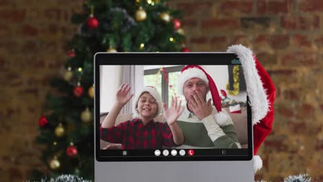 Happy-father-and-son-waving-on-video-call-on-computer,-with-christmas-decorations-and-tree