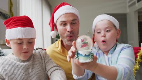 Kaukasischer-Vater-Und-Zwei-Söhne-Mit-Weihnachtsmützen-Und-Einer-Schneekugel,-Die-Zu-Hause-Auf-Der-Couch-Sitzen