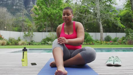Happy-african-american-plus-size-woman-practicing-yoga,-stretching-next-to-swimming-pool-in-garden