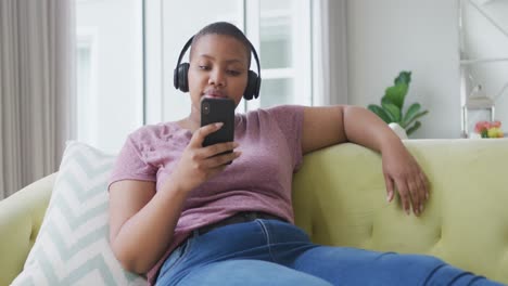 Happy-african-american-plus-size-woman-sitting-on-sofa,-wearing-headphones-and-using-smartphone