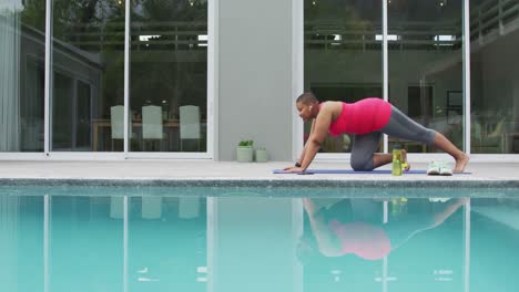 Happy-african-american-plus-size-woman-practicing-yoga,-stretching-next-to-swimming-pool-in-garden