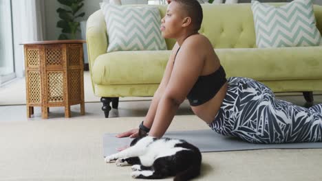 Feliz-Mujer-Afroamericana-De-Talla-Grande-Practicando-Yoga,-Estirándose-En-La-Sala-De-Estar-Con-Su-Gato-Mascota