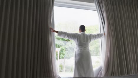 Happy-african-american-plus-size-woman-wearing-robe,-drawing-curtains-and-looking-through-window