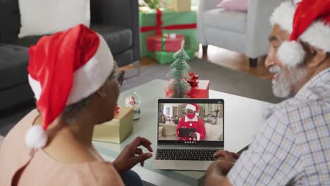 Happy-african-american-couple-on-video-call-on-laptop-with-santa-claus-in-face-mask-at-christmas
