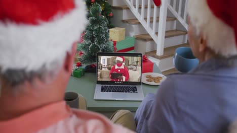 Happy-senior-caucasian-couple-on-video-call-on-laptop-with-santa-in-face-mask-at-christmas-time
