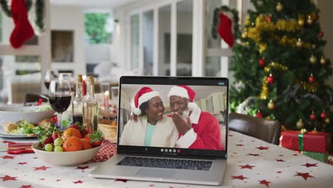 Happy-african-american-senior-couple-in-santa-hats-on-laptop-lying-on-christmas-table