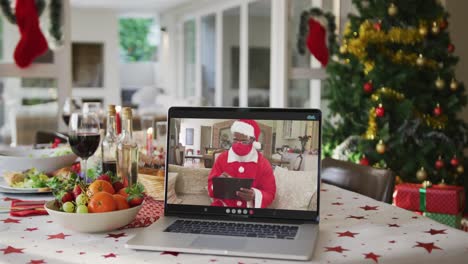 Glücklicher-Afroamerikanischer-Weihnachtsmann-Mit-Gesichtsmaske-Auf-Laptop,-Der-Auf-Dem-Weihnachtstisch-Liegt