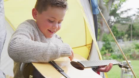 Happy-caucasian-boy-sitting-in-tent-in-garden-and-playing-guitar