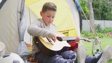 Caucasian-boy-sitting-in-tent-in-garden-and-playing-guitar