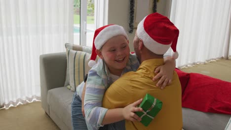 Caucasian-boy-wearing-santa-hat-holding-a-christmas-gift-hugging-his-father-at-home