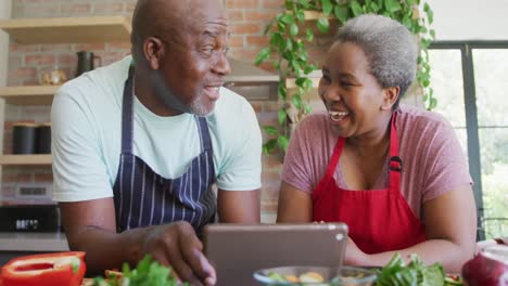 Glückliches-Afroamerikanisches-Seniorenpaar,-Das-Zusammen-Kocht-Und-Ein-Tablet-In-Der-Küche-Verwendet