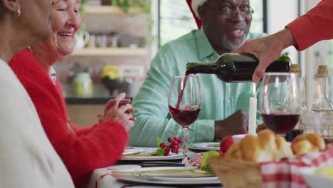 Happy-diverse-female-and-male-senior-friends-celebrating-meal,-pouring-vine-at-christmas-time