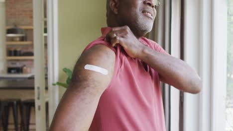 Portrait-of-african-american-senior-man-showing-his-vaccinated-shoulder-at-home