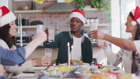 Happy-group-of-diverse-friends-in-santa-hats-celebrating-meal,-toasting-with-juice-at-christmas-time