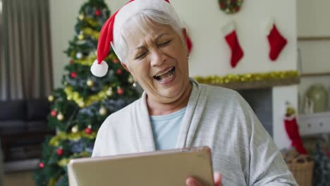 Happy-mixed-race-senior-woman-in-santa-hat-on-video-call-on-tablet-at-christmas-time