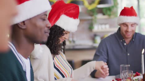 Happy-group-of-diverse-friends-in-santa-hats-praying,-celebrating-meal-at-christmas-time