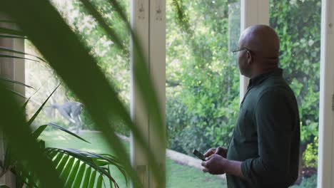 Afroamerikanischer-älterer-Mann,-Der-Sein-Smartphone-Benutzt,-Während-Er-Zu-Hause-Aus-Dem-Fenster-Schaut