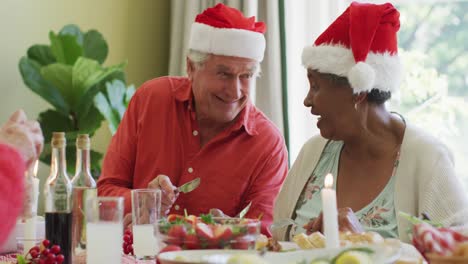 Fröhliches,-Vielfältiges-älteres-Paar-Mit-Weihnachtsmützen,-Das-Zu-Hause-Am-Weihnachtstisch-Lacht