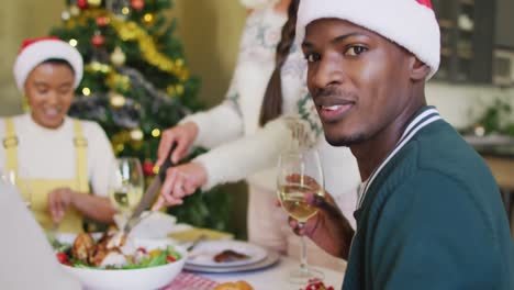 Happy-african-american-man-in-santa-hat-celebrating-meal-with-friends-at-christmas-time