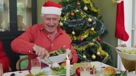 Happy-caucasian-senior-man-in-santa-hat-serving-himself-food,-at-christmas-dinner-table-with-friends
