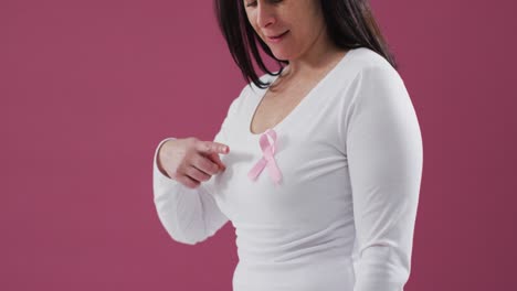 Mid-section-of-a-woman-pointing-to-the-pink-ribbon-on-her-chest-against-pink-background