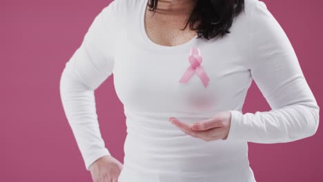 Mid-section-of-woman-with-pink-ribbon-on-her-chest-holding-invisible-object-against-pink-background