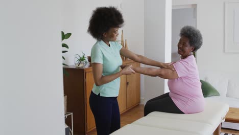 African-american-female-physiotherapist-wearing-face-mask-helping-senior-female-patient-exercise