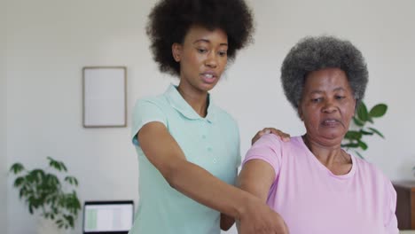 Happy-african-american-female-physiotherapist-helping-senior-female-patient-exercise-at-home