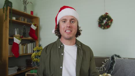 Glücklicher-Kaukasischer-Mann-Mit-Weihnachtsmütze-Bei-Einem-Weihnachtsvideoanruf,-Der-Ein-Geschenk-In-Der-Hand-Hält-Und-Redet