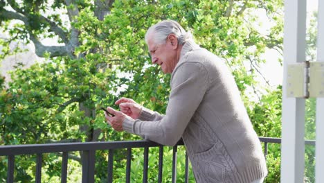 Side-view-of-senior-man-leaning-on-railing-while-using-smart-phone-in-balcony
