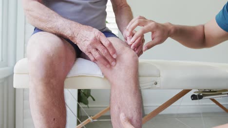 Male-physiotherapist-examining-knee-joint-of-senior-man-on-examination-table-at-studio