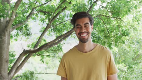 Portrait-of-cheerful-biracial-man-in-yellow-t-shirt-is-standing-against-trees
