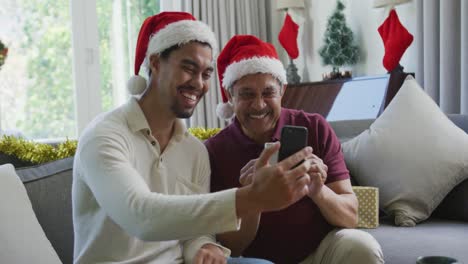 Happy-biracial-son-with-father-in-santa-hats-doing-video-call-on-smartphone-in-christmas