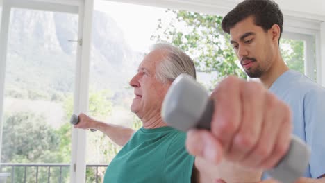 Male-physiotherapist-looking-at-senior-man-exercising-with-dumbbells-in-retirement-home