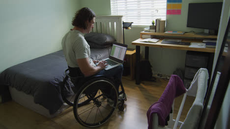 Fokussierter-Kaukasischer-Behinderter-Mann-Im-Rollstuhl,-Der-Laptop-Im-Schlafzimmer-Benutzt