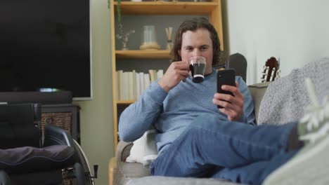 Happy-caucasian-disabled-man-sitting-on-sofa-in-living-room-using-smartphone-and-drinking-coffee