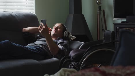 Relaxed-caucasian-disabled-man-lying-on-sofa-in-living-room-wearing-headphones-and-using-smartphone