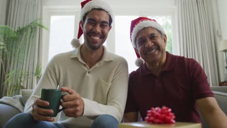 Portrait-of-happy-biracial-man-and-father-in-santa-hats-making-christmas-video-call-at-home