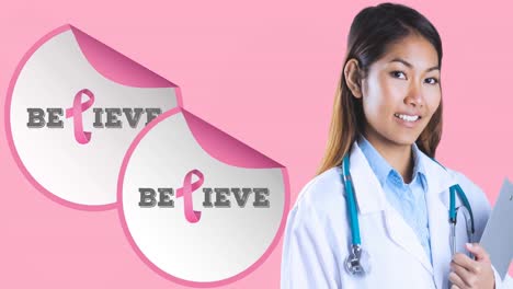 Animation-of-believe-text-with-pink-ribbon-over-smiling-biracial-female-doctor-on-pink-background