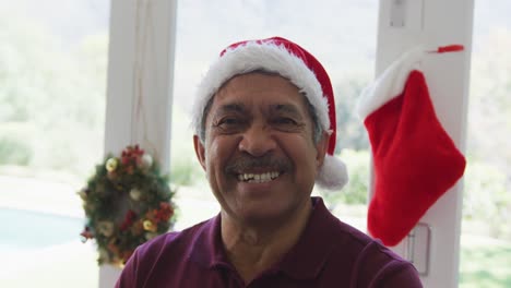 Portrait-of-cheerful-senior-man-in-santa-hat-at-decorated-home-during-christmas